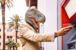 Senior Mann tragen T-Rex Dinosaurier Maske abheben Geld von Bank Kasse Maschine mit Lastschrift Karte - - surreal Bild von Hälfte Mensch und Tier - - absurd und verrückt Konzept von Geldautomat werben foto