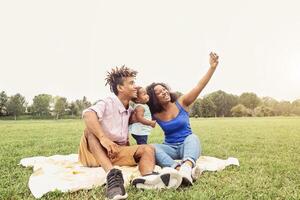 glücklich afrikanisch Familie tun Selfie Foto mit Handy, Mobiltelefon Telefon im ein Öffentlichkeit Park draussen - - Mutter und Vater haben Spaß mit ihr Tochter während ein Wochenende sonnig Tag - - Liebe und Glück Konzept
