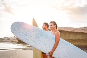 jung Paar von glücklich Surfer Stehen auf das Strand halten Surfbretter vorbereiten zu Surfen auf hoch Wellen während ein großartig Sonnenuntergang - - Menschen, Lebensstil, extrem Sport Konzept - - Fokus auf Mann foto