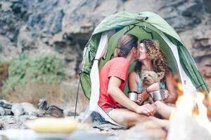 Reise Paar Camping im Felsen Berge nach ein Trekking Tag - - glücklich Menschen Trinken heiß Tee Innerhalb Zelt mit ihr Hund Nächster zu Lagerfeuer - - Sport, Beziehung, Liebe und Ferien Konzept foto