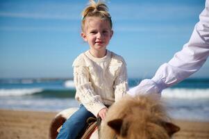Nahansicht Porträt von glücklich Kind Mädchen Reiten ein Pferd Pony auf das Strand, lächelnd suchen beim Kamera foto