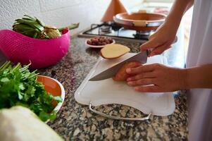 Nahansicht Aussicht Frau Hände mit Küche Messer, Hacken organisch Kartoffel auf ein Schneiden Planke, Kochen Abendessen beim Zuhause Küche foto