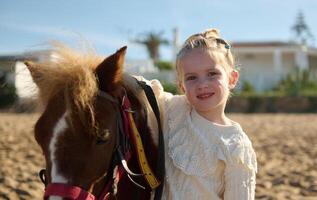 bezaubernd kaukasisch Kind Mädchen umarmen ein wenig Pony, lächelnd suchen beim Kamera, genießen ein glücklich Familie Wochenende draussen. foto