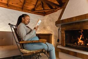 Senior afrikanisch Frau lesen Buch während Erwärmen im Vorderseite von Kamin im ihr Haus foto