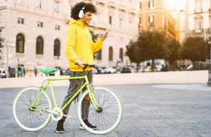 glücklich afro Mann mit Handy, Mobiltelefon Smartphone draussen - - jung Kerl haben Spaß Hören Musik- mit Kopfhörer während Reiten mit Fahrrad im Stadt - - Jugend tausendjährig Generation Lebensstil und Technologie Konzept foto