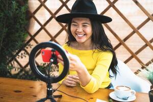 glücklich asiatisch Frau vlogging online mit Smartphone Nocken und Influencer LED während Trinken ein Kaffee Sitzung im Bar foto