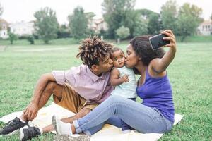 glücklich afrikanisch Familie nehmen ein Selfie mit Handy, Mobiltelefon Clever Telefon Kamera im ein Öffentlichkeit Park draussen - - Mutter und Vater haben Spaß mit ihr Tochter während ein Wochenende sonnig Tag - - Liebe und Glück Konzept foto