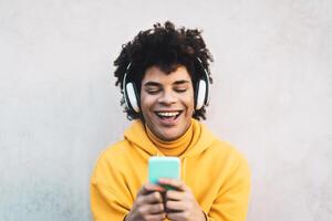 glücklich afro Mann mit Handy, Mobiltelefon Smartphone draussen - - jung Kerl haben Spaß Hören Musik- mit kabellos Kopfhörer draussen - - Jugend tausendjährig Generation Lebensstil und Menschen süchtig Technologie Konzept foto