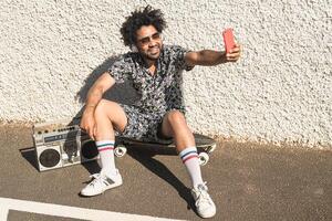 jung afro Latein Mann haben Spaß nehmen Selfie mit Handy, Mobiltelefon Smartphone während Hören Musik- mit Kopfhörer und Jahrgang Boombox während Sommer- Urlaube foto