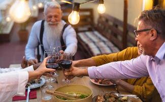 glücklich gemischtrassig Senior freunde Toasten mit rot Wein Brille zusammen auf Haus Terrasse Abendessen - - Alten Lebensstil Menschen und Essen Konzept foto