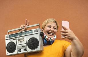 Plus Größe Frau Hören zu Musik- mit Jahrgang Boombox Stereo während nehmen Selfie mit Handy, Mobiltelefon Smartphone während Corona Virus Pandemie foto