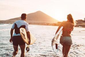 glücklich Surfer Paar Laufen mit Surfbretter entlang das Meer Ufer - - sportlich Menschen haben Spaß gehen zu Surfen zusammen beim Sonnenuntergang - - extrem Surfen Sport und Jugend Beziehung Lebensstil Konzept foto