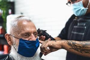 männlich Friseur Schneiden Haar zu Hipster Senior Klient während tragen Gesicht chirurgisch Maske - - jung Friseur Arbeiten im Friseur während Corona Virus Ausbruch - - Gesundheit Pflege und Haarschnitt Salon Konzept foto