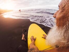 glücklich Surfer Senior Sitzung auf Surfbrett Aufpassen Sonnenuntergang Zeit - - reifen bärtig Mann haben Spaß auf Surfen Tag - - extrem Sport und Gesundheit Menschen Lebensstil Konzept foto