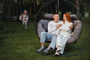 ein glücklich Familie ist Sitzung im ein Hängematte auf das Rasen in der Nähe von das Haus foto