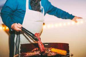 Senior Kochen Fleisch auf Grill - - Koch Grillen Fleisch und Portion Würste beim Abendessen auf Dach - - Konzept von Essen Grill und Essen zusammen draussen - - Fokus auf Mann Hand foto