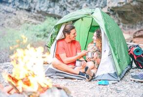 glücklich Paar von Wanderer mit ihr Hund Camping mit Zelt um Felsen Berge Nächster zu Feuer - - Menschen entspannend im ein Lager Trinken heiß Tee nach ein Trekking Tag - - reisen, Liebe, Sport und Lebensstil Konzept foto