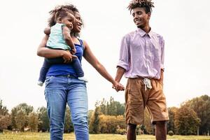 glücklich afrikanisch Familie Gehen zusammen im Öffentlichkeit Park - - Mutter, Vater und Baby Tochter haben Spaß während ein Wochenende sonnig Tag draussen - - Glück, Liebe und Elternschaft Konzept foto