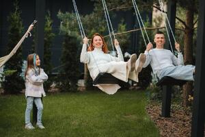 Mama und Papa sind Reiten auf ein schwingen und Dort ist ein Tochter Stehen Nächster zu ihnen. das Familie ist ruhen auf ein schwingen foto