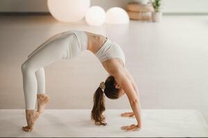 ein Mädchen im Weiß Kleider tut Yoga Stehen auf das Brücke auf ein Matte drinnen foto