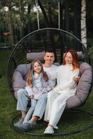 ein glücklich Familie ist Sitzung im ein Hängematte auf das Rasen in der Nähe von das Haus foto