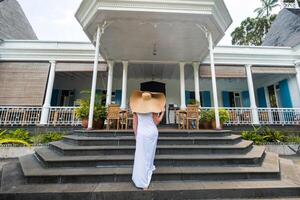 das Frau im das groß Hut ist lächelnd. ein schön Mädchen im ein groß Hut und Weiß Kleid lächelt draußen ein alt kolonial Gebäude auf das Insel von Mauritius foto