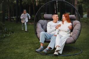 ein glücklich Familie ist Sitzung im ein Hängematte auf das Rasen in der Nähe von das Haus foto