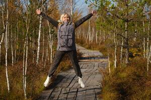 ein glücklich Frau mit ein Rucksack springt auf ein hölzern Pfad im ein Sumpf im Yelnya, Weißrussland foto