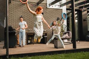 ein glücklich Familie ist ruhen nicht weit von ihr heim. Papa ist Kochen ein Grill, und Mama und Tochter sind spielen Springen foto