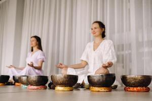 zwei Frauen sind Sitzung mit tibetanisch Schalen im das Lotus Position Vor ein Yoga Klasse im das Fitnessstudio foto