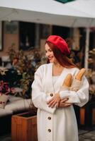 Porträt von ein ziemlich Frau im ein rot Baskenmütze und ein Weiß Strickjacke mit Baguettes im ihr Hände spazieren gehen durch das Herbst Stadt foto