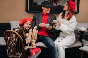 ein stilvoll Familie von drei ist Sitzung beim ein Tabelle draußen im ein Cafe und Trinken Kaffee. Papa, Mama und Tochter im das Herbst Stadt foto