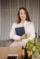 Porträt von ein lächelnd Mädchen Frau Sitzung im ein Sessel. ein Aromatherapeut im ein Weiß Bluse ist Sitzung im das Büro foto