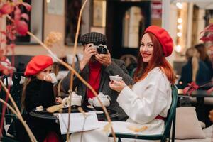 ein stilvoll Familie von drei ist Sitzung beim ein Tabelle draußen im ein Cafe und Trinken Kaffee. Papa nimmt Bilder von Mama im das Herbst Stadt foto