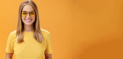 stilvoll selbstbewusst charmant europäisch Frau mit Messe Haar und Braun gebrannt Haut im Gelb T-Shirt und Sonnenbrille lächelnd breit amüsiert und heiter Blick beim Kamera posieren gegen Orange Hintergrund foto