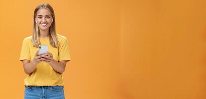 Porträt von freundlich charismatisch und abgehend gut aussehend jung weiblich im Gelb T-Shirt halten Smartphone tragen kabellos Kopfhörer genießen Hören Musik- auf Weg zu Fitness Fitnessstudio foto