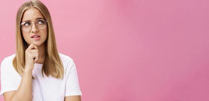 Studio Schuss von unsicher Clever nerdig Frau im Brille und Weiß T-Shirt Stehen beunruhigt und besorgt berühren Kinn suchen gestört beim Oberer, höher richtig Ecke zögernd, Denken gegen Rosa Mauer foto