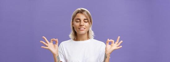 friedlich Ruhe gut aussehend weiblich blond im Weiß T-Shirt Schließen Augen lächelnd erleichtert und glücklich Gefühl zufrieden mit Leben Stehen im Lotus Pose mit Zen Geste Über lila Hintergrund foto