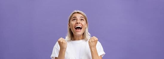 hüfthoch Schuss von dankbar Erfreut Glücklich Mädchen mit attraktiv bräunen im Weiß T-Shirt Zusammenpressen Faust von positiv Emotionen Blick Erfreut und glücklich oben dankbar geben Vielen Dank Gott zum erfüllen Traum foto