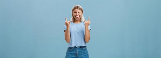 Lassen uns Felsen diese Party. Porträt von froh gut aussehend und sorglos jung künstlerisch weiblich Musiker mit blond Haar zeigen schwer Metall Geste kleben aus Zunge und zwinkert amüsiert, haben Spaß foto