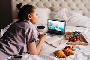 ein Mädchen sitzt im Bett im das Abend, mit ein Smartphone im ihr Hand und isst Erdbeeren, ein Mädchen im Bett hat Süßigkeiten Vor gehen zu Bett. foto