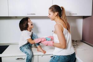 Hausfrau Mama im Rosa Handschuhe wäscht Geschirr mit ihr Sohn durch Hand im das sinken mit Waschmittel. ein Mädchen im Weiß und ein Kind mit ein Besetzung reinigt das Haus und wäscht Geschirr im hausgemacht Rosa Handschuhe. foto