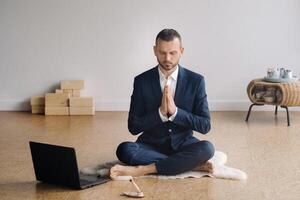 ein Mann im ein formal passen meditiert während Sitzung im ein Fitness Zimmer mit ein Laptop foto