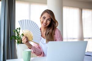 ein jung Frau schwitzt reichlich während Arbeiten auf ihr Laptop im ein überhitzt Zuhause Büro. mit Nein Luft Conditioner, sie haftet zu ein Welle Fan, kämpfen das heftig Sommer- Hitze. foto