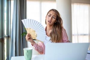 ein jung Frau schwitzt reichlich während Arbeiten auf ihr Laptop im ein überhitzt Zuhause Büro. mit Nein Luft Conditioner, sie haftet zu ein Welle Fan, kämpfen das heftig Sommer- Hitze. foto