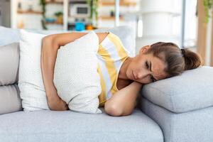 Frau, die auf dem Sofa liegt und im Wohnzimmer krank aussieht. schöne junge frau, die auf bett liegt und hände auf ihrem bauch hält. frau mit schmerzhaften bauchschmerzen im bett, menstruationsperiode foto