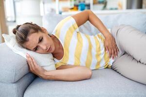 Frau, die auf dem Sofa liegt und im Wohnzimmer krank aussieht. schöne junge frau, die auf bett liegt und hände auf ihrem bauch hält. frau mit schmerzhaften bauchschmerzen im bett, menstruationsperiode foto