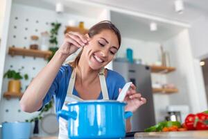 froh jung Dame Kochen und Probenahme Abendessen im ein Topf, Geschenk im ein zeitgenössisch Zuhause Küche. Hausfrau vorbereiten nahrhaft Mahlzeit mit ein lächeln. inländisch Leben und Nahrung. gesund Essen. foto