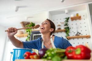 komisch schön Frau Singen in Spatel, Kochen im modern Küche, halten Spatel wie Mikrofon, Tanzen, Hören zu Musik, spielerisch Mädchen haben Spaß mit Geschirr, vorbereiten Lebensmittel. foto