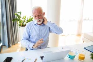 Nackenschmerzen. Senior Mann berühren Schmerz Hals Leiden von Schmerzen Sitzung Arbeiten auf Laptop im modern Büro. Gesundheitspflege, Gesundheit Probleme im älter Alter Konzept foto